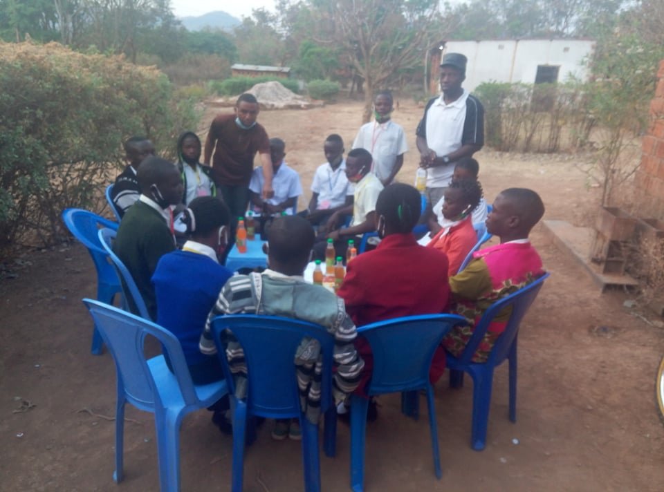 Group of children and adults gathered around a blue outdoor table with drinks, some children wearing face masks, in a rural outdoor setting.