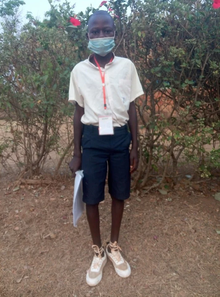 A young boy standing outdoors in front of bushes, wearing a face mask, a light-colored shirt, dark shorts, tan shoes, and holding a piece of paper.