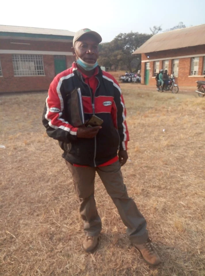 A man standing outdoors on a dry grassy ground, wearing a red and black jacket, brown pants, and a tan cap. He holds a notebook and a smartphone. In the background, there are brick buildings, trees, and people on bicycles or motorcycles.