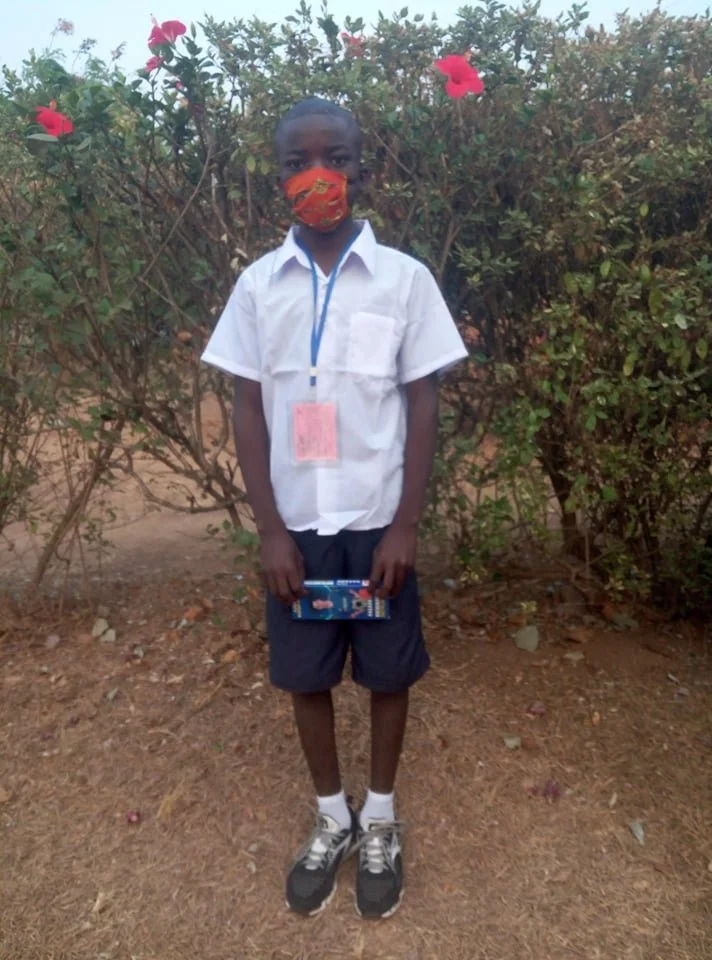 A young boy in a school uniform, wearing a face mask and standing on dirt ground in front of bushes with pink flowers, holding a book or notebook.