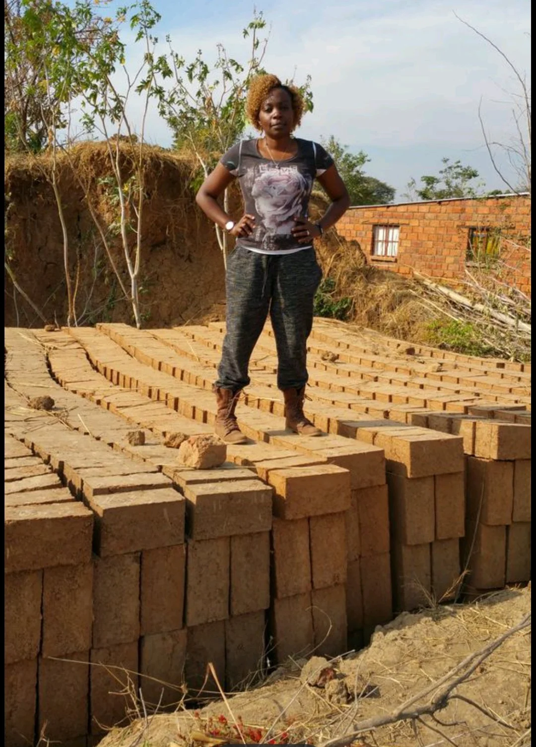 Woman standing on a partially constructed brick wall outdoors, with trees and a brick building in the background.