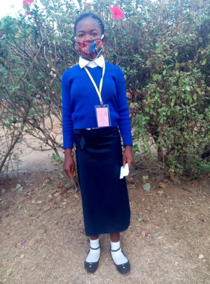 A girl wearing a blue sweater, white shirt, and a long black skirt stands outdoors in front of bushes with pink flowers, wearing a face mask and a yellow lanyard with an ID badge.