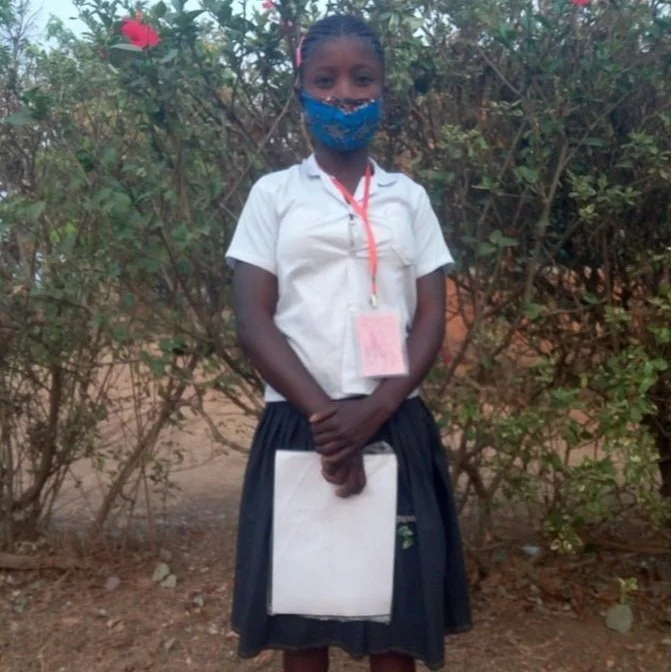A young girl in a school uniform, wearing a blue face mask, standing outdoors in front of bushes, holding a notebook.
