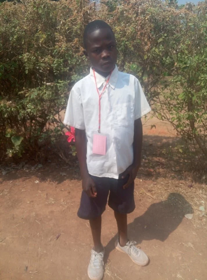 A young boy in a white school shirt and dark shorts standing on dirt ground outdoors. He is wearing a pink ID badge around his neck and has a serious expression. There are green bushes in the background.