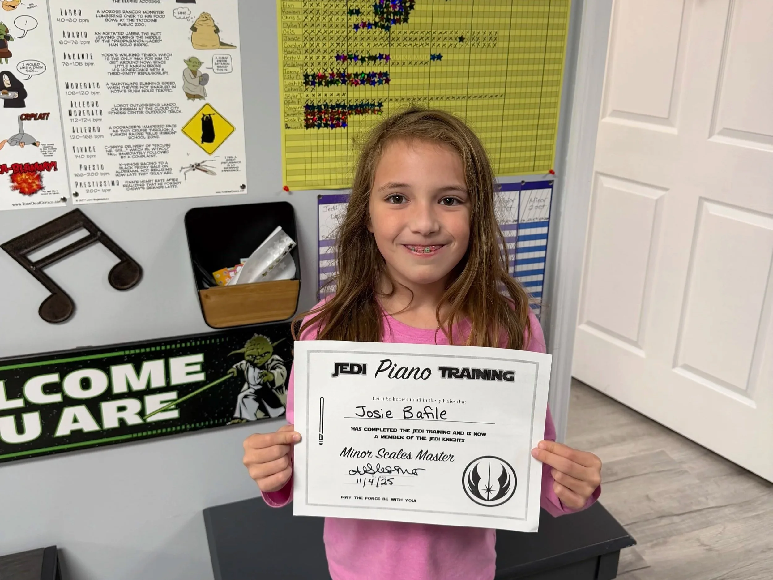 A young girl with long hair and a pink shirt holding a Jedi Jedi Training certificate in a classroom. Behind her are educational posters, charts, and a Star Wars-themed welcome sign.