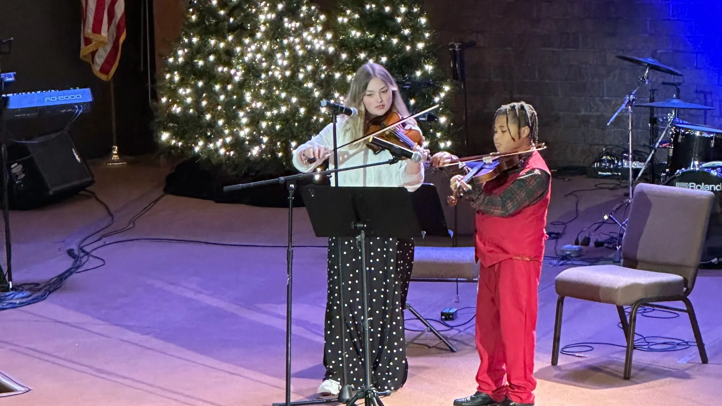 Two young girls playing violins on stage during a Christmas performance, with a lit Christmas tree in the background and musical equipment around them.