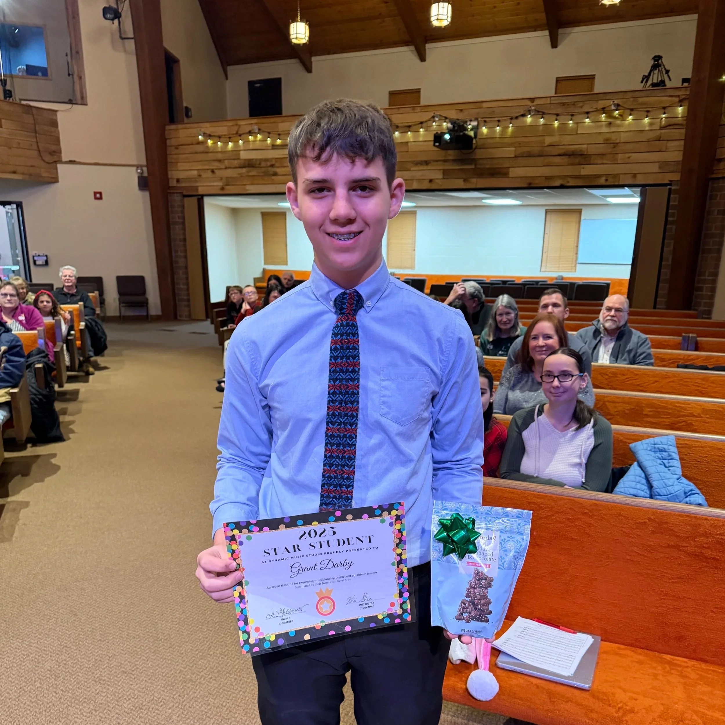 A young boy in a blue shirt and patterned tie standing in a church, holding a certificate, a gift bag with a green bow, a bag of chocolate, and a Santa hat, with an audience of seated adults and children behind him.