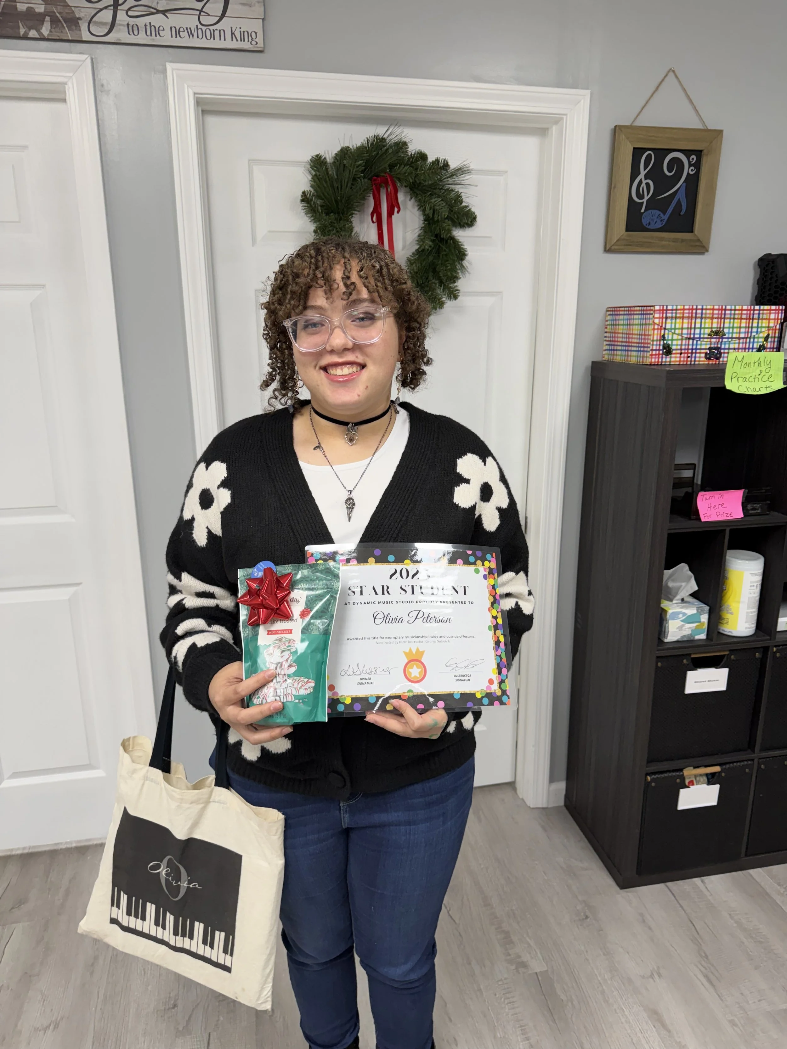 A young woman smiling and holding a certificate of achievement, a gift bag, and a wrapped present. She stands indoors, wearing glasses, a necklaces, a black and white sweater, and jeans. There is a Christmas wreath on the door behind her and various signs and shelves in the background.