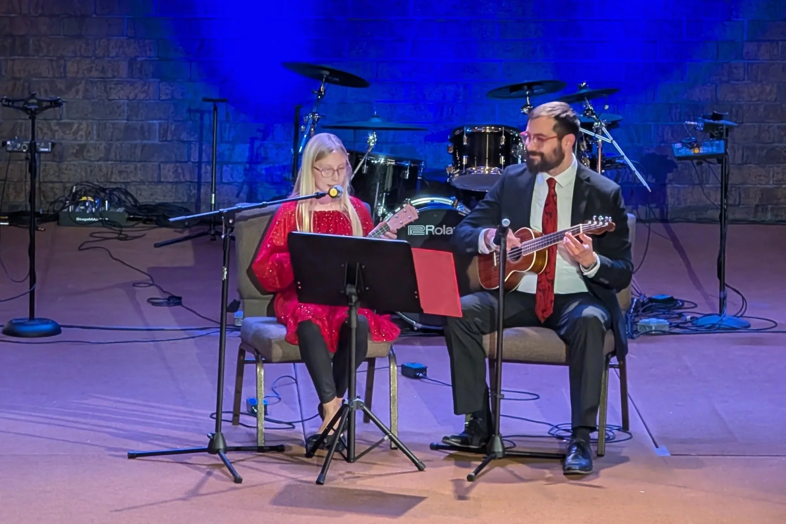 A young girl and an adult man are sitting on chairs on a stage, playing ukuleles and singing into microphones. The girl is wearing a red dress and glasses, and the man is dressed in a dark suit with a red tie. In the background, there is a drum set on a brick wall.