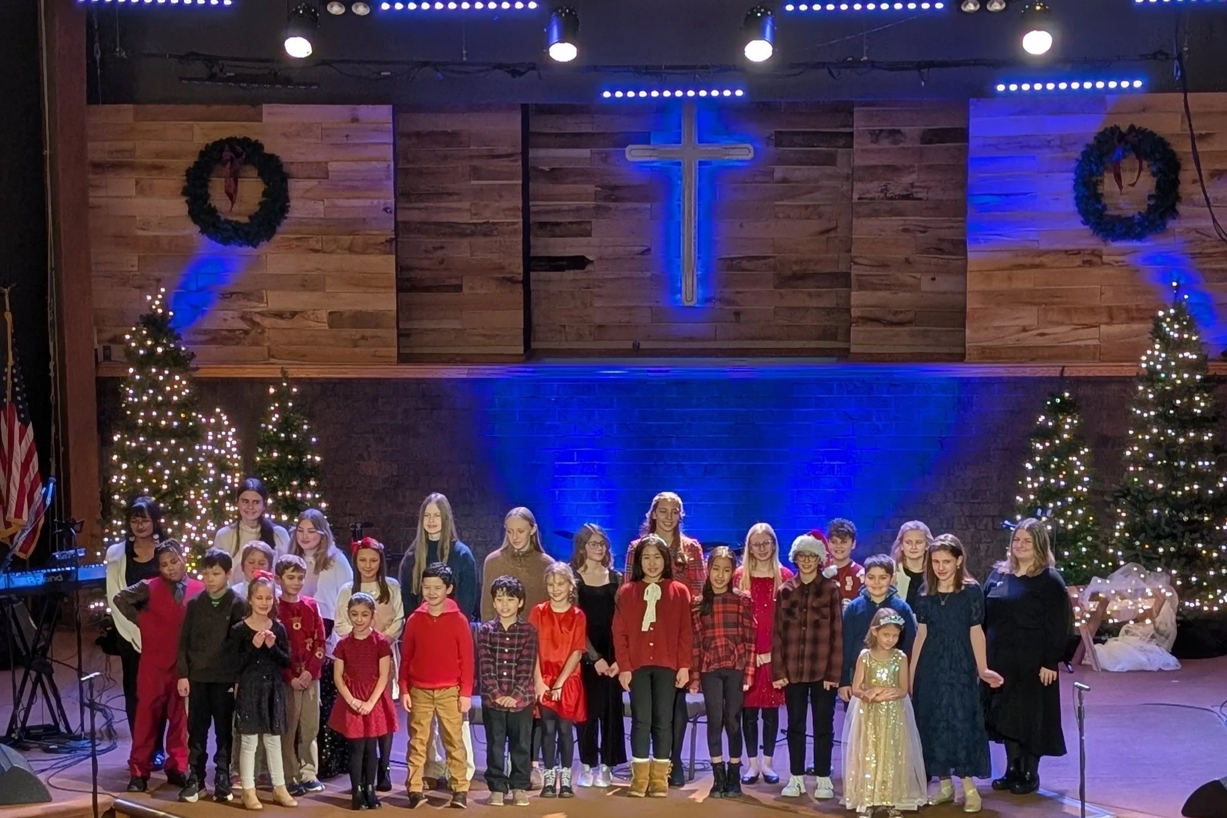 A group of children and adults standing on stage in front of Christmas trees and a cross in a church, decorated for Christmas.