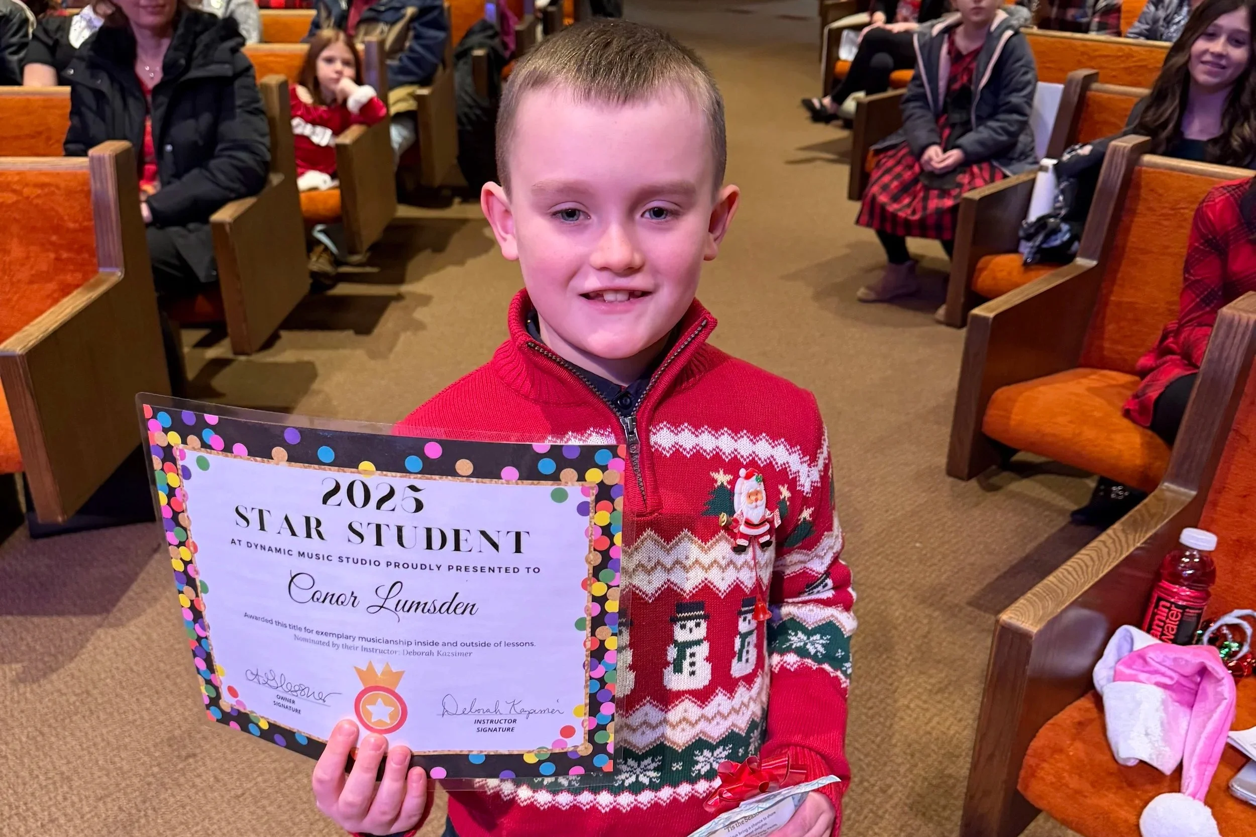A young boy in a red Christmas sweater with snowman designs holding a certificate for 2025 Star Student at a music studio, smiling in a church or auditorium with other children seated in the background.