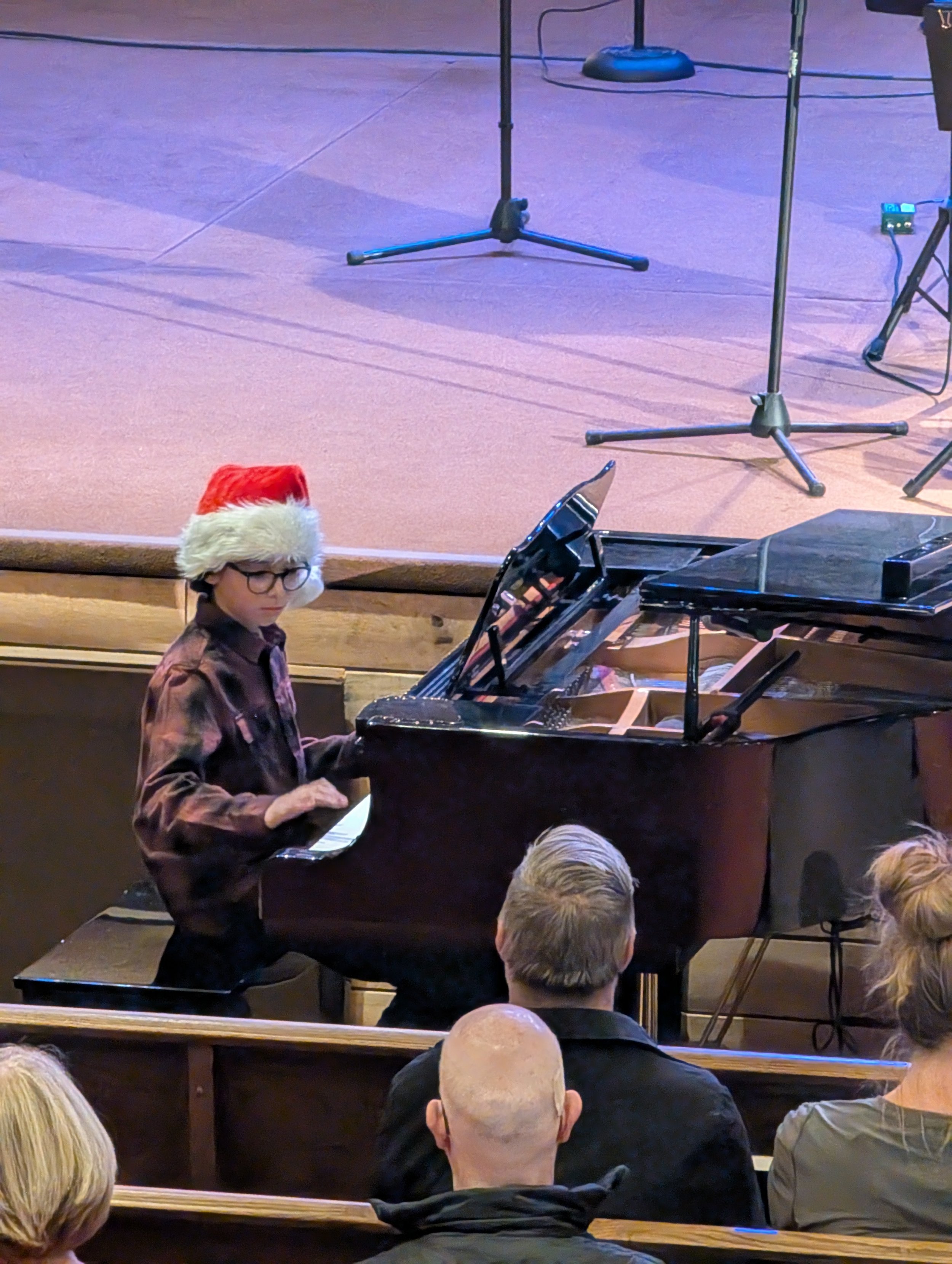 A boy wearing a Santa hat and glasses playing the piano during a holiday performance in front of an audience.