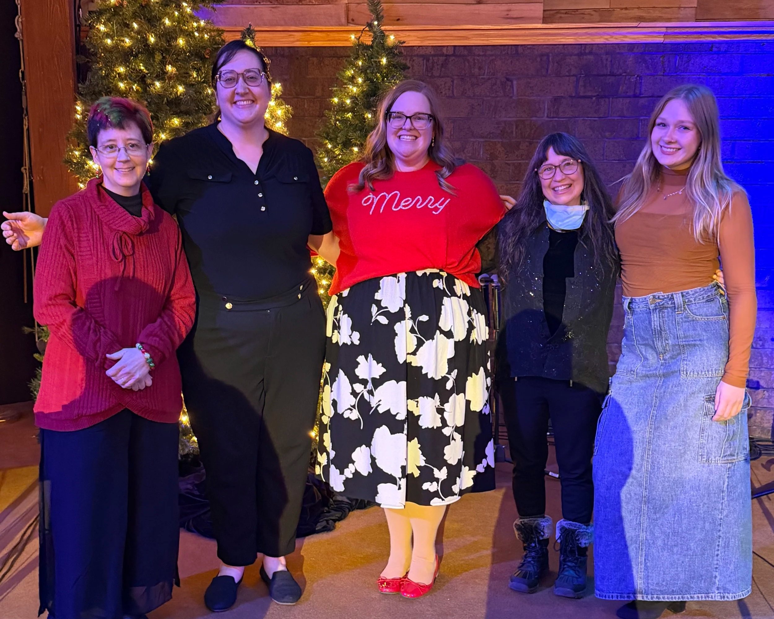 Six women standing in front of Christmas trees decorated with lights. They are posing with smiles and dressed in festive and casual clothing, celebrating the holiday season.