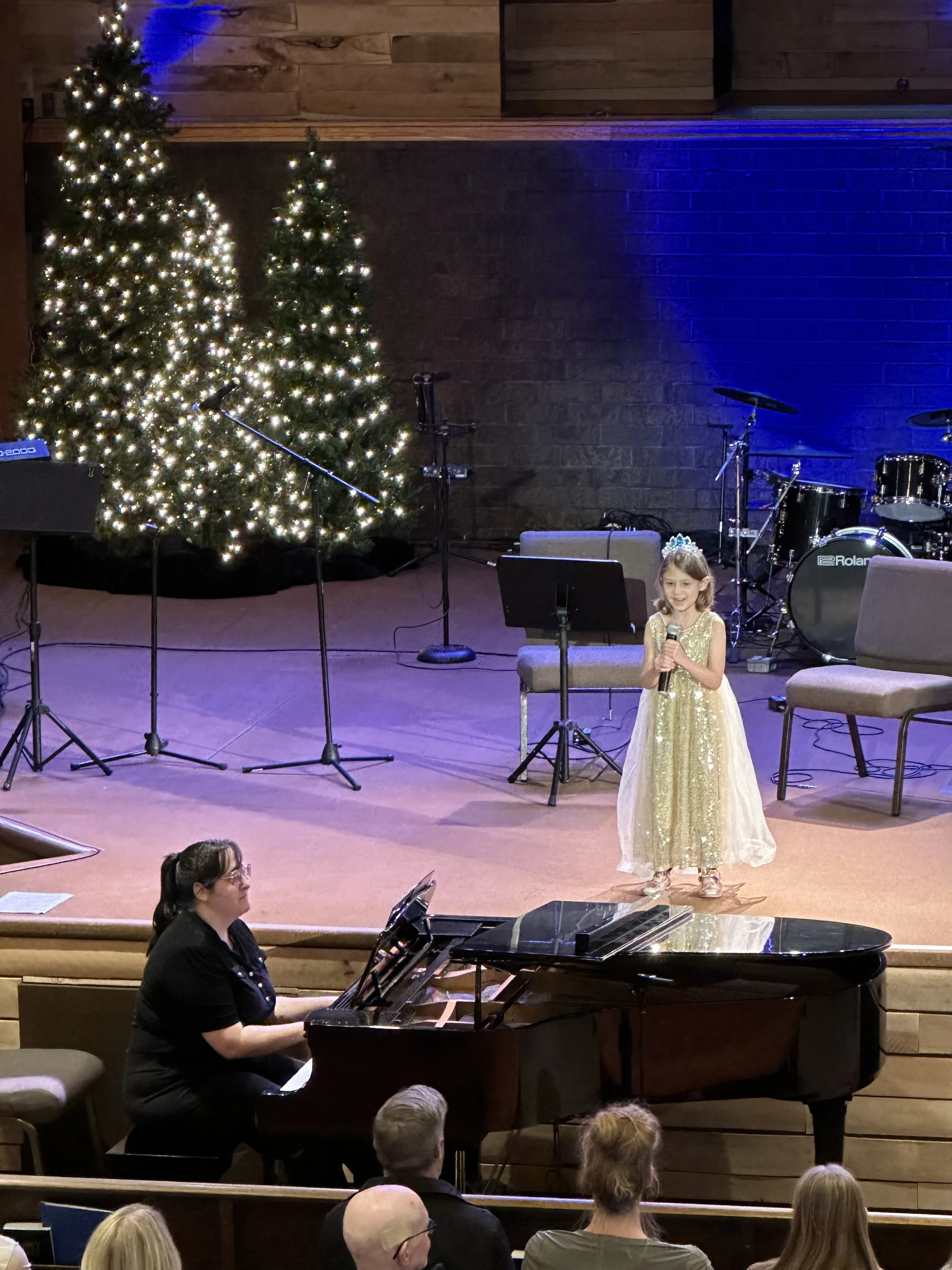 Young girl in a gold dress and tiara singing into a microphone on stage, accompanied by a woman playing grand piano, with Christmas trees decorated with white lights and musical instruments in the background.