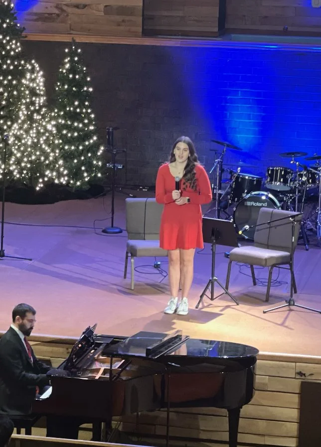 A woman in a red dress holding a microphone stands on a stage with Christmas trees decorated with lights behind her. There is a music stand, two empty chairs, and a man playing a grand piano in front of the stage. The background features a brick wall illuminated with blue lighting.