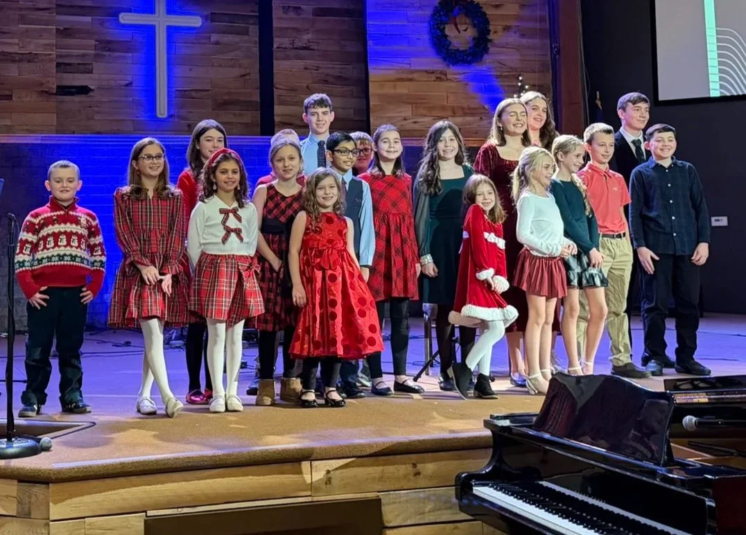 Children dressed in Christmas-themed clothing standing on a stage with a cross and wreath in the background, singing or performing.