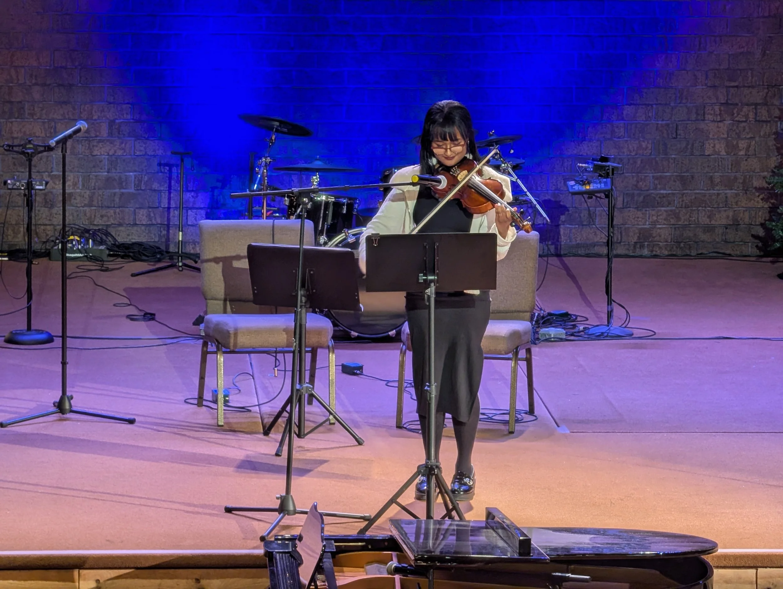 A woman performing on stage with a violin, music stand, and microphone, wearing a dark dress and glasses, with drums and musical equipment in the background.