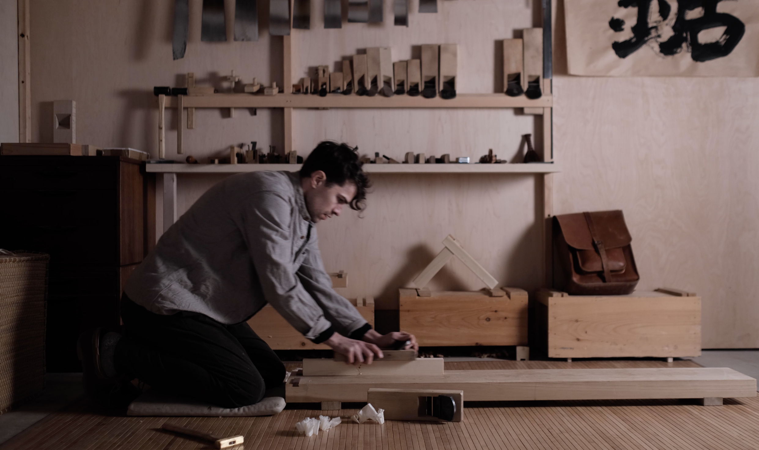 A man kneeling on a rug, working with a wooden board in a woodworking shop. Shelves with various woodworking tools and materials are seen in the background.