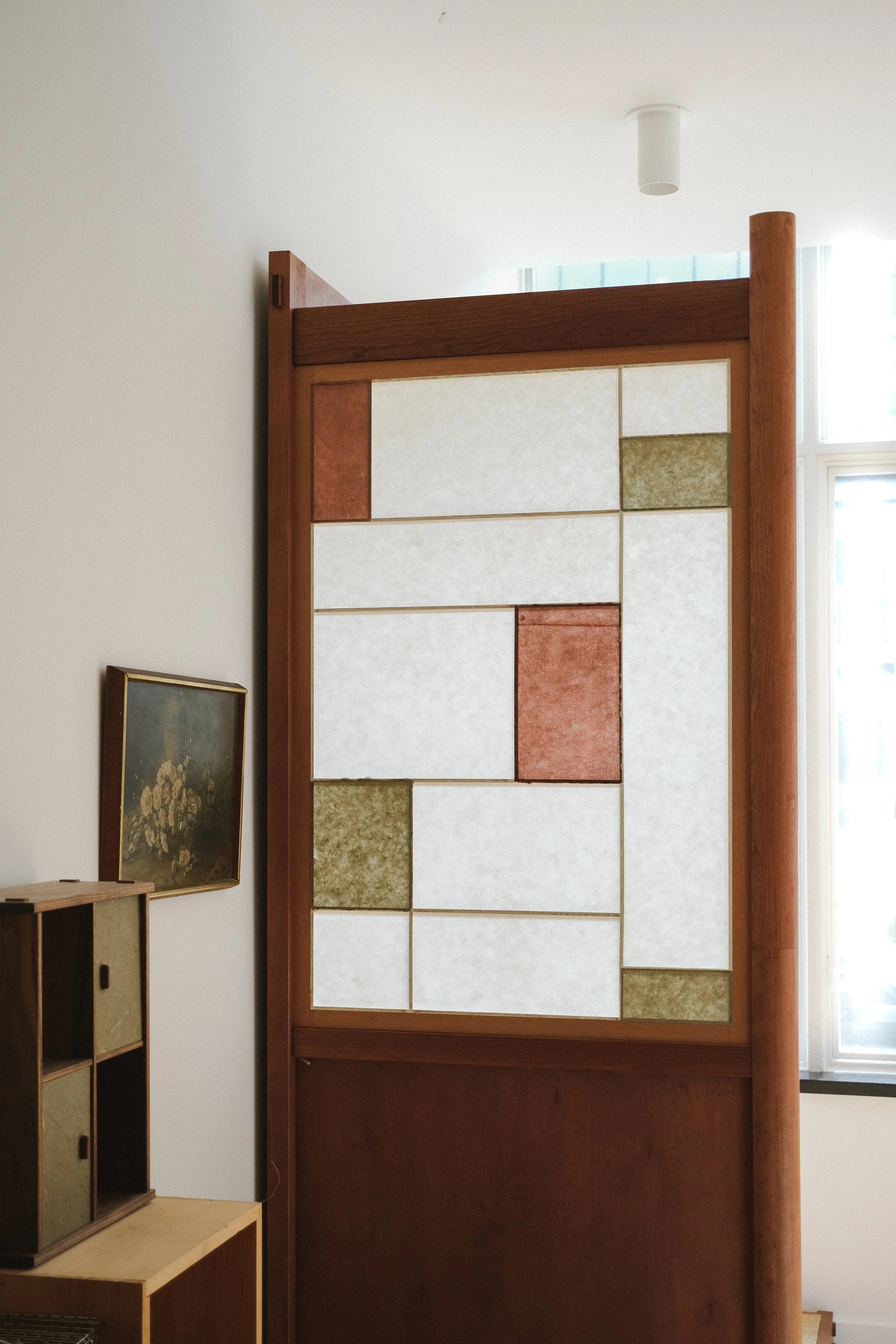 Interior of a room with a decorative wooden room divider featuring geometric panels in white, green, and red, positioned near a window with sheer curtains. Part of a wooden shelving unit and a framed floral painting are visible.