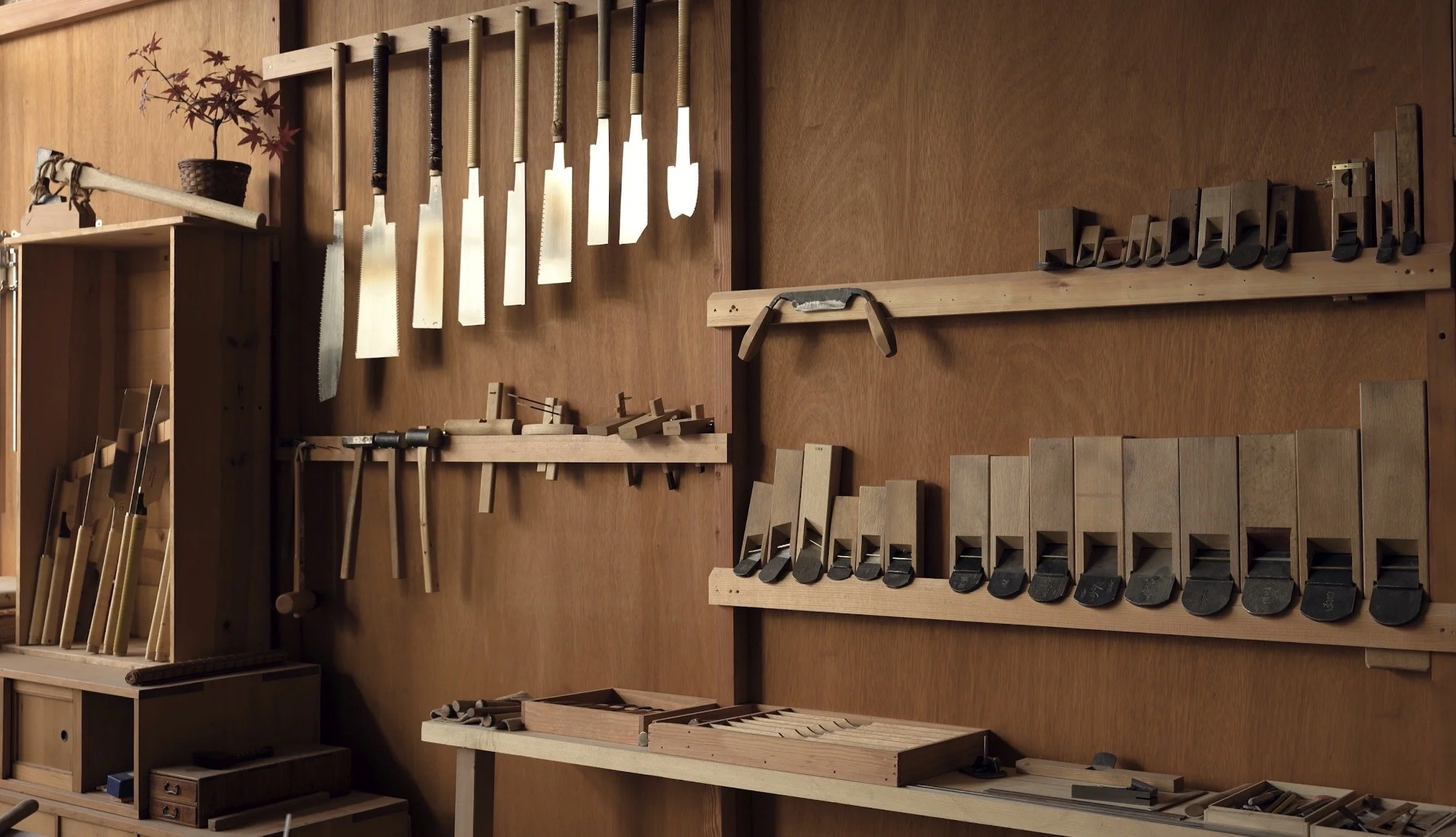 A woodworking shop wall with various hand tools including saws, chisels, and planes organized on wooden shelves and hanging on the wall.