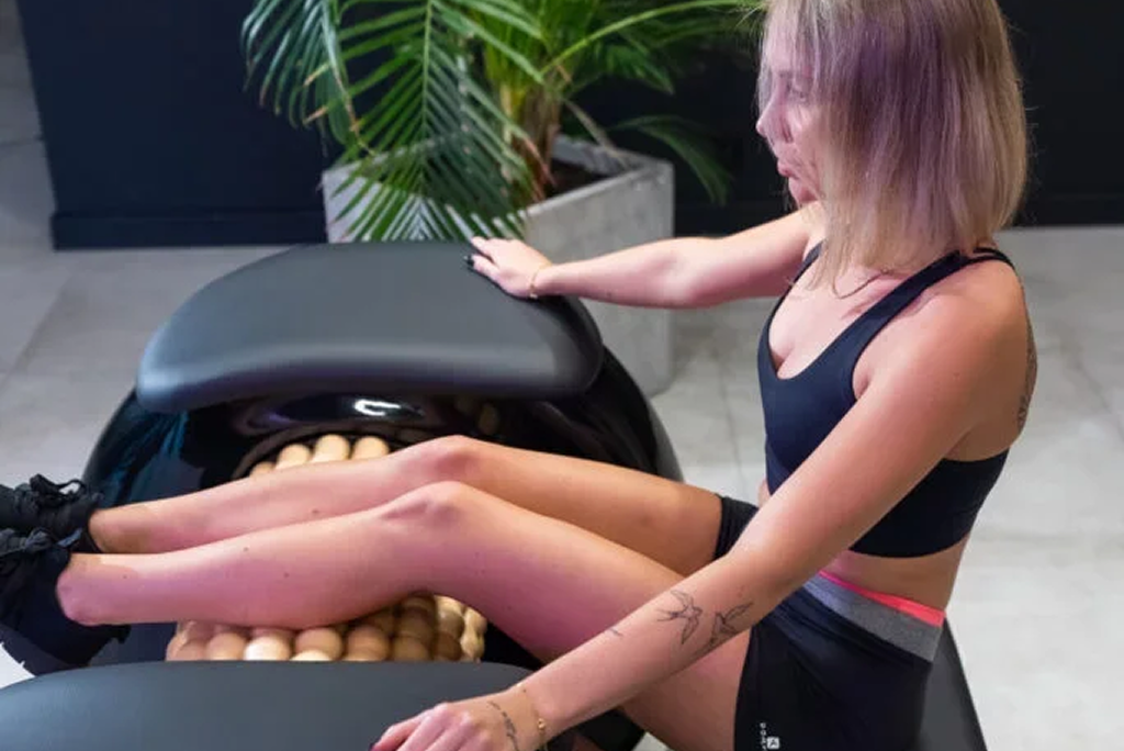 A woman with short blonde hair in a black sports bra and black shorts is sitting on a massage roller machine with her legs extended. She has a tattoo on her left forearm and is holding the machine's surface while looking down.