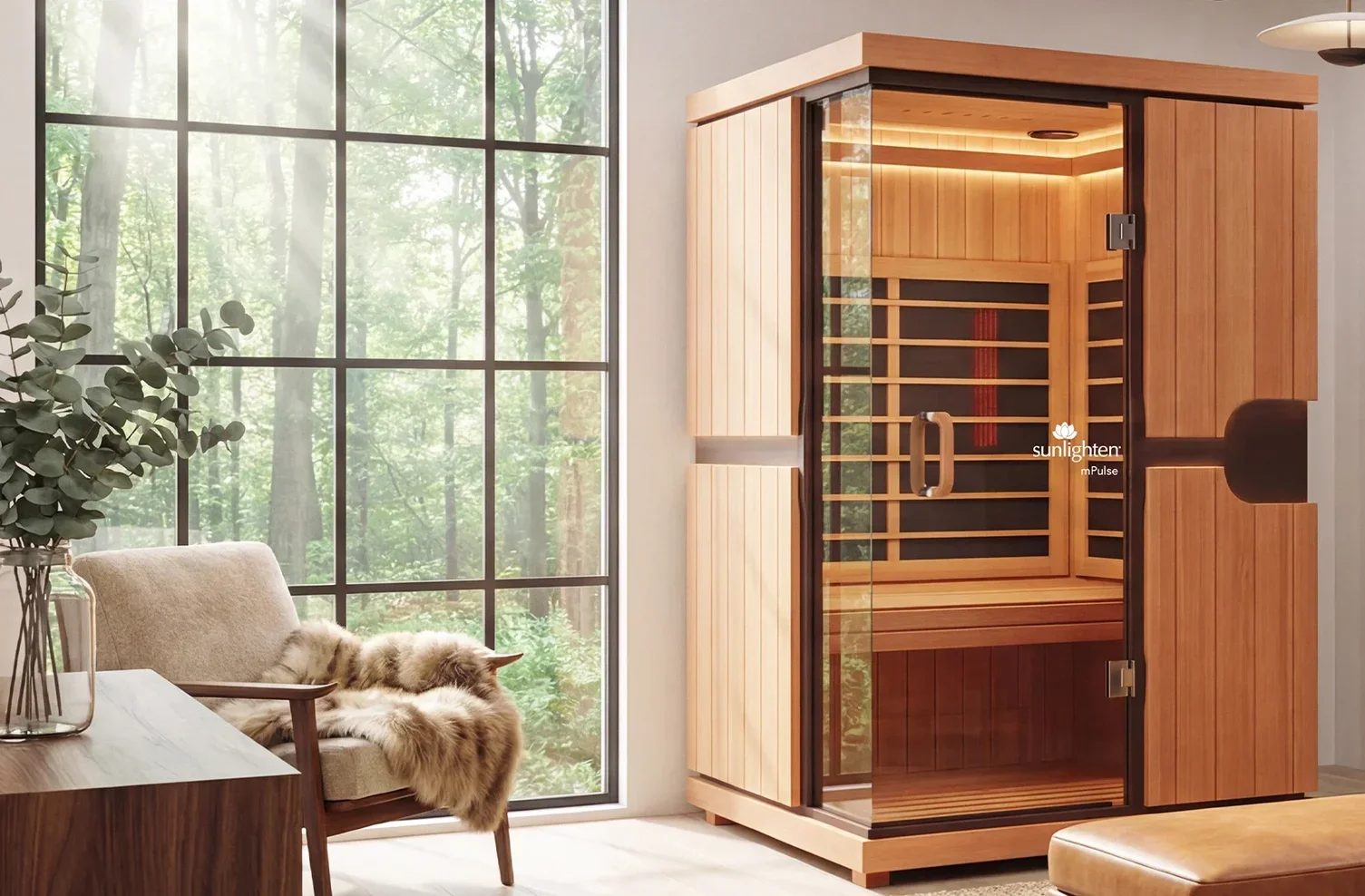 A modern interior room with large window showing a forest outside, a beige armchair draped with a fur throw, a wooden side table with a glass vase of eucalyptus, and a freestanding wooden infrared sauna.