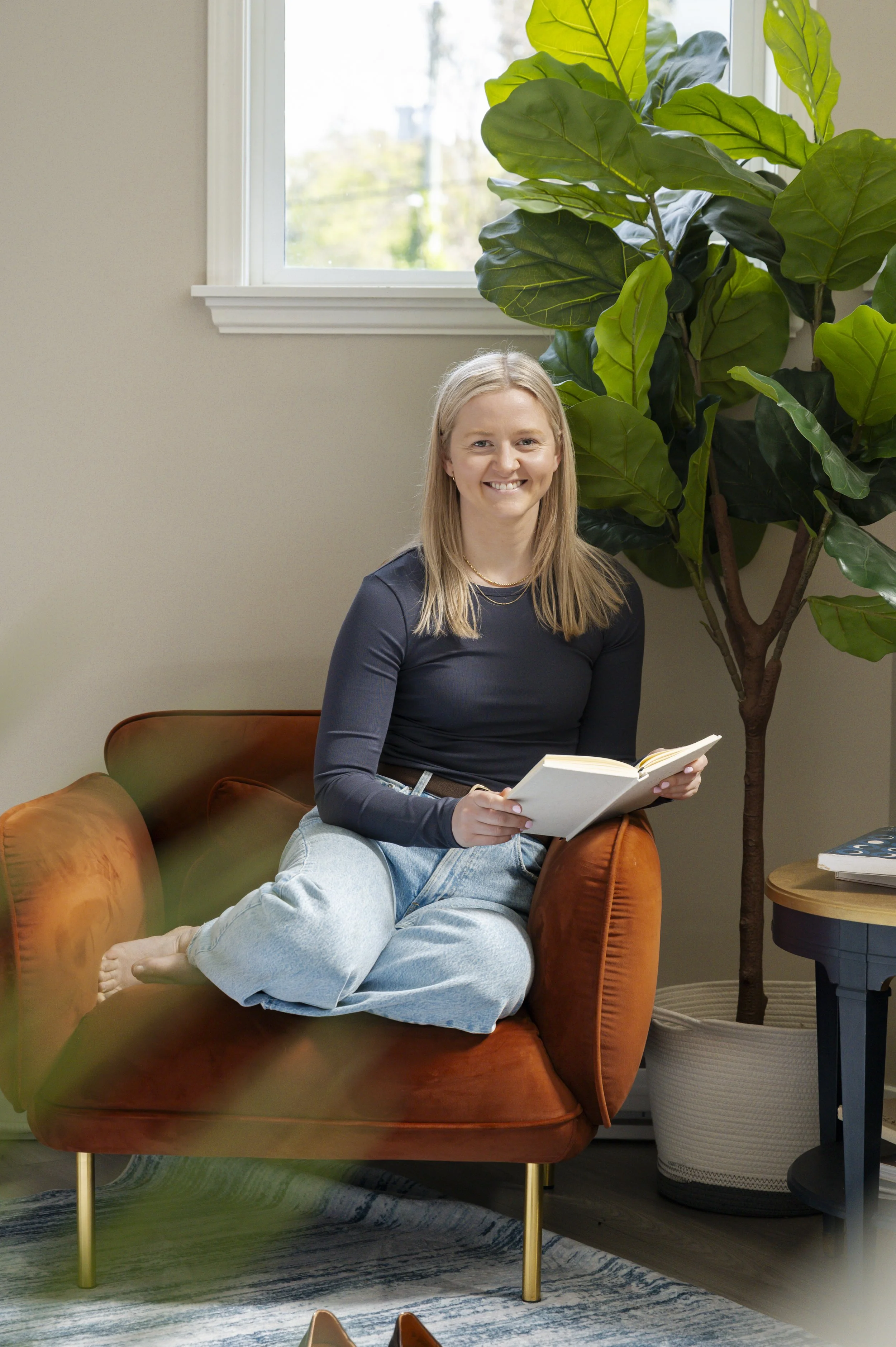 A woman with blonde hair, smiling, sitting on a brown velvet armchair, reading a book. She is wearing a black long-sleeve top and light blue jeans, with her legs crossed and barefoot. There is a large green houseplant to her right and a white wall with a window behind her. There is a small round side table with books on it next to the armchair.