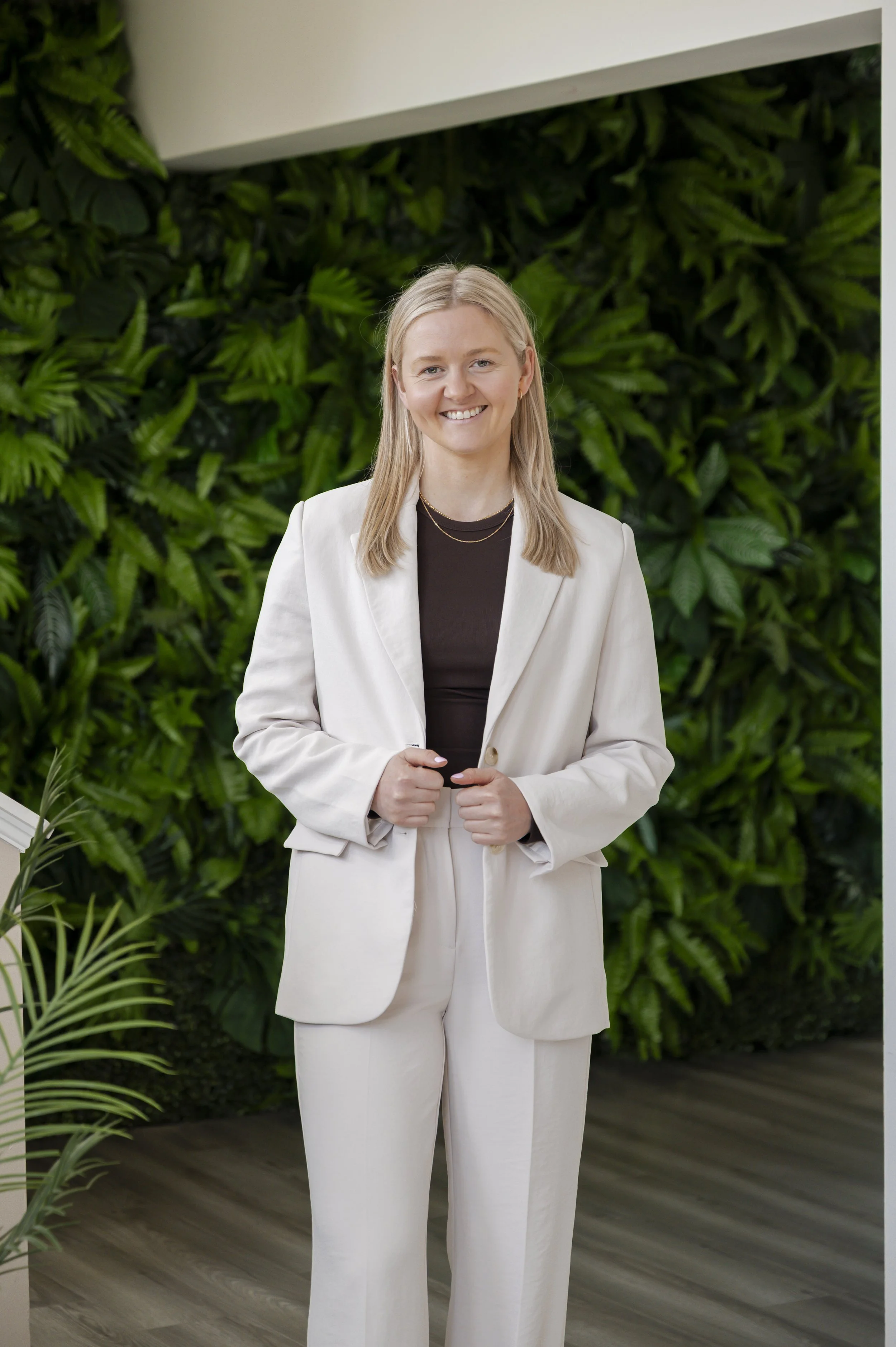 A woman with long blonde hair smiling, dressed in a white suit with a black top, standing in front of a green wall of plants inside a building.