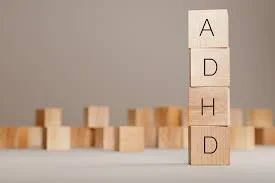 Four wooden blocks stacked vertically spelling 'ADHD' beside a row of small wooden blocks on a gray surface.