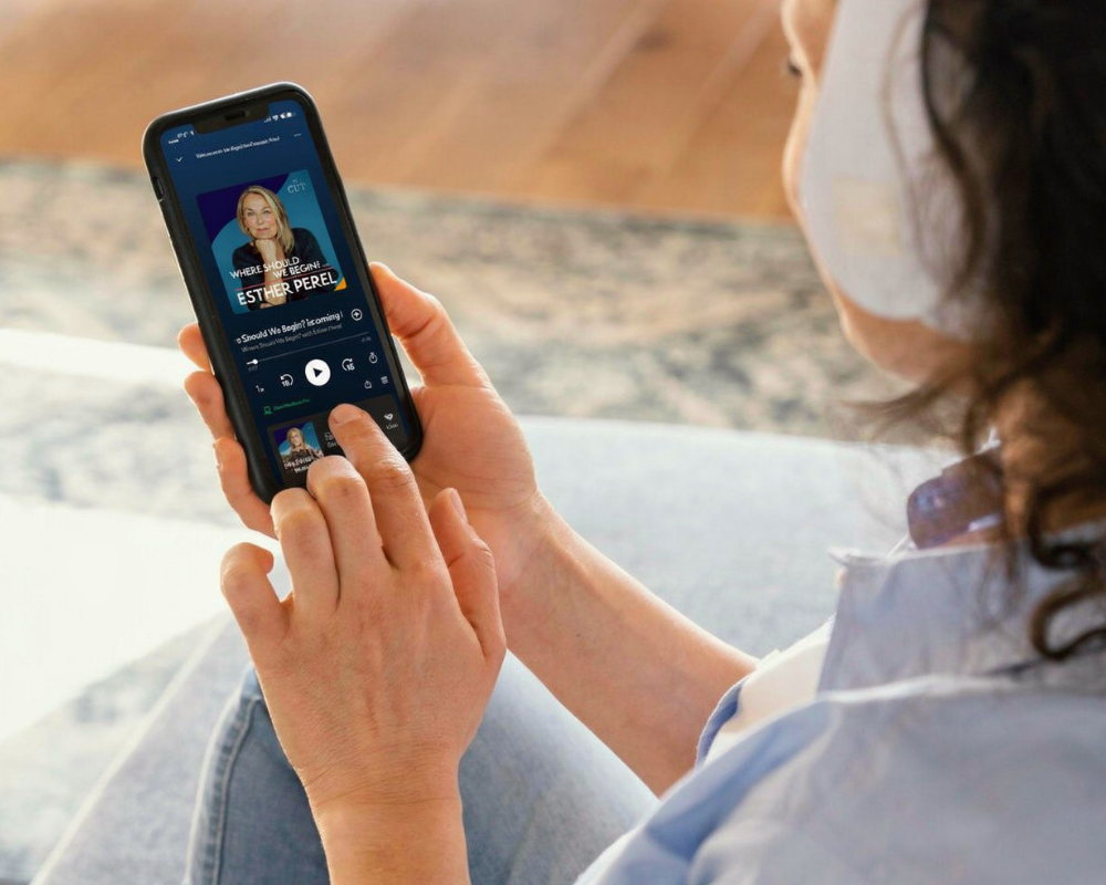 A woman with curly hair and a face mask is sitting on a couch, holding a smartphone and browsing a podcast app.