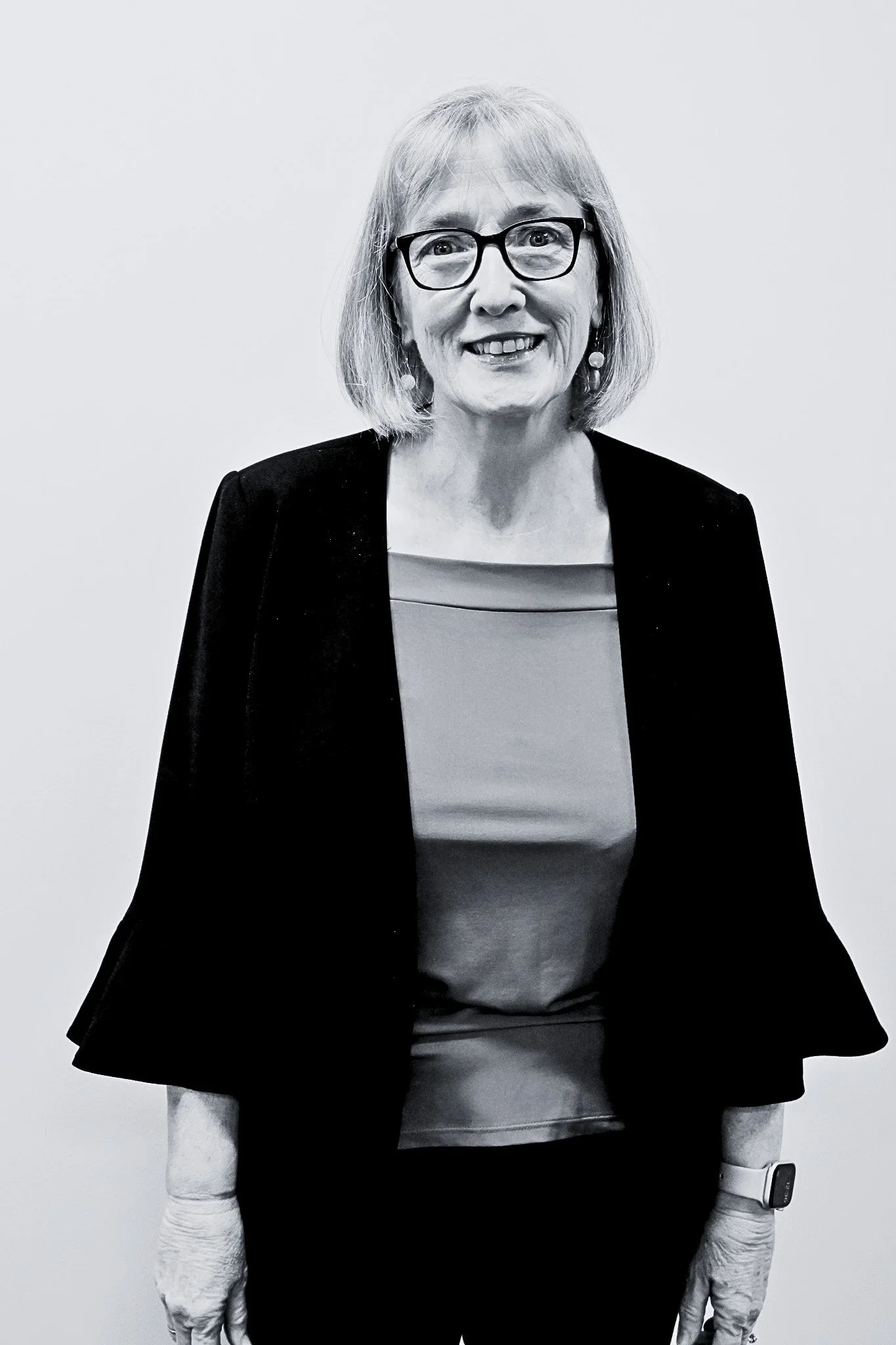 A black and white photograph of a middle-aged woman with glasses, wearing a black jacket, a top, and gloves. She is smiling and looking directly at the camera, standing against a plain background.