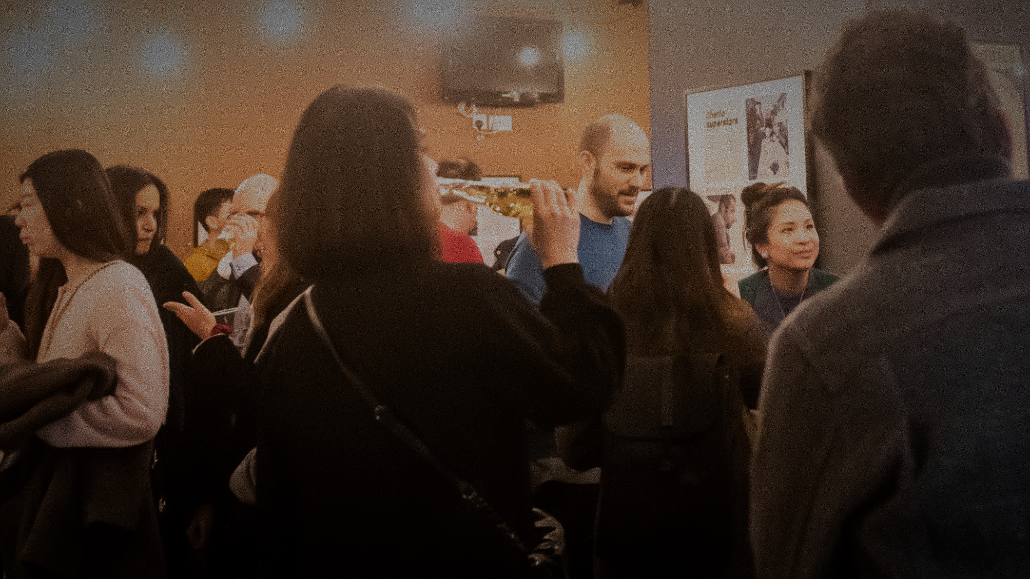 A group of people gathered indoors at a social or networking event. The setting has warm lighting and a casual atmosphere. Some attendees are engaged in conversation, while others appear to be holding drinks or snacks.