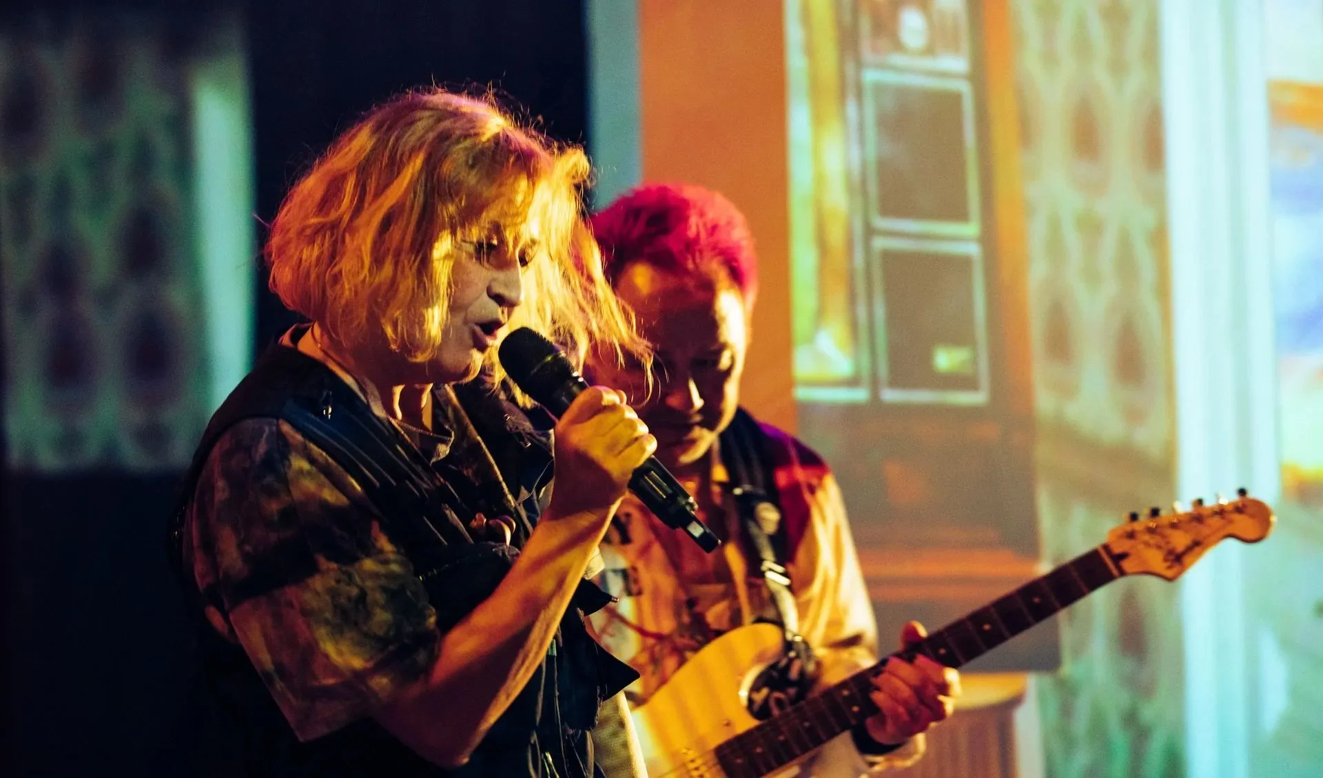 A woman sings into a microphone on stage while a man plays the guitar behind her
