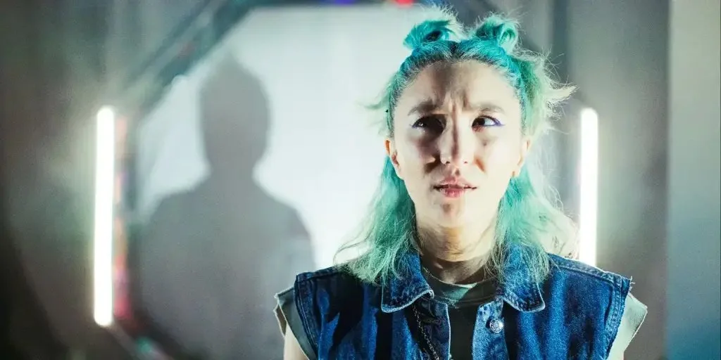 A woman with green hair and wearing a sleevless, denim jacket faces forward with a confused expression