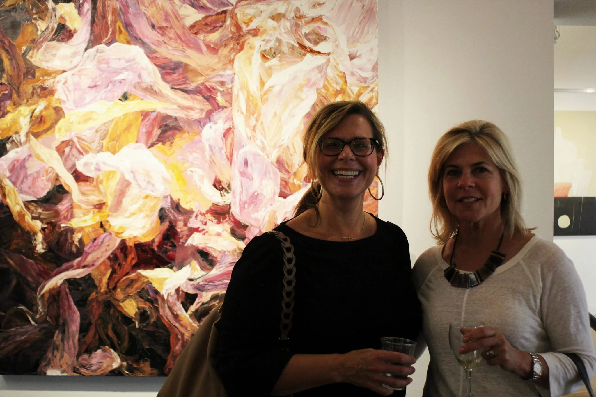 Two women are standing in front of an abstract painting at an art gallery, holding drinks and smiling at the camera.