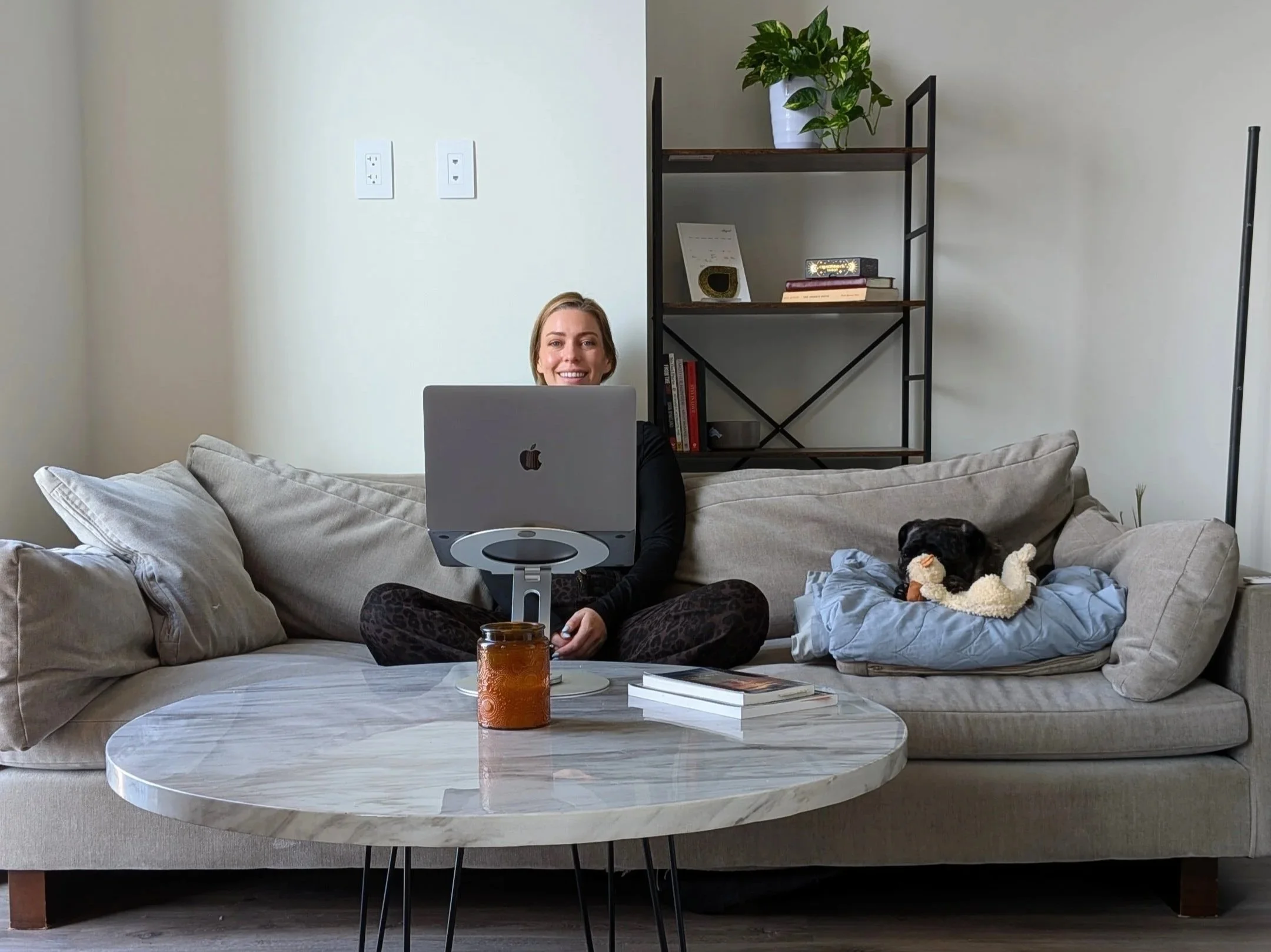 A woman is sitting on a beige couch in a modern living room, smiling while working on a silver Apple laptop. There is a small black dog lying on a blue blanket beside her. In front of her is a white marble coffee table with an orange candle and a few books. Behind her is a black metal and wood bookshelf with a potted plant, a framed certificate, and several books. The room has light-colored walls with two electrical outlets visible.