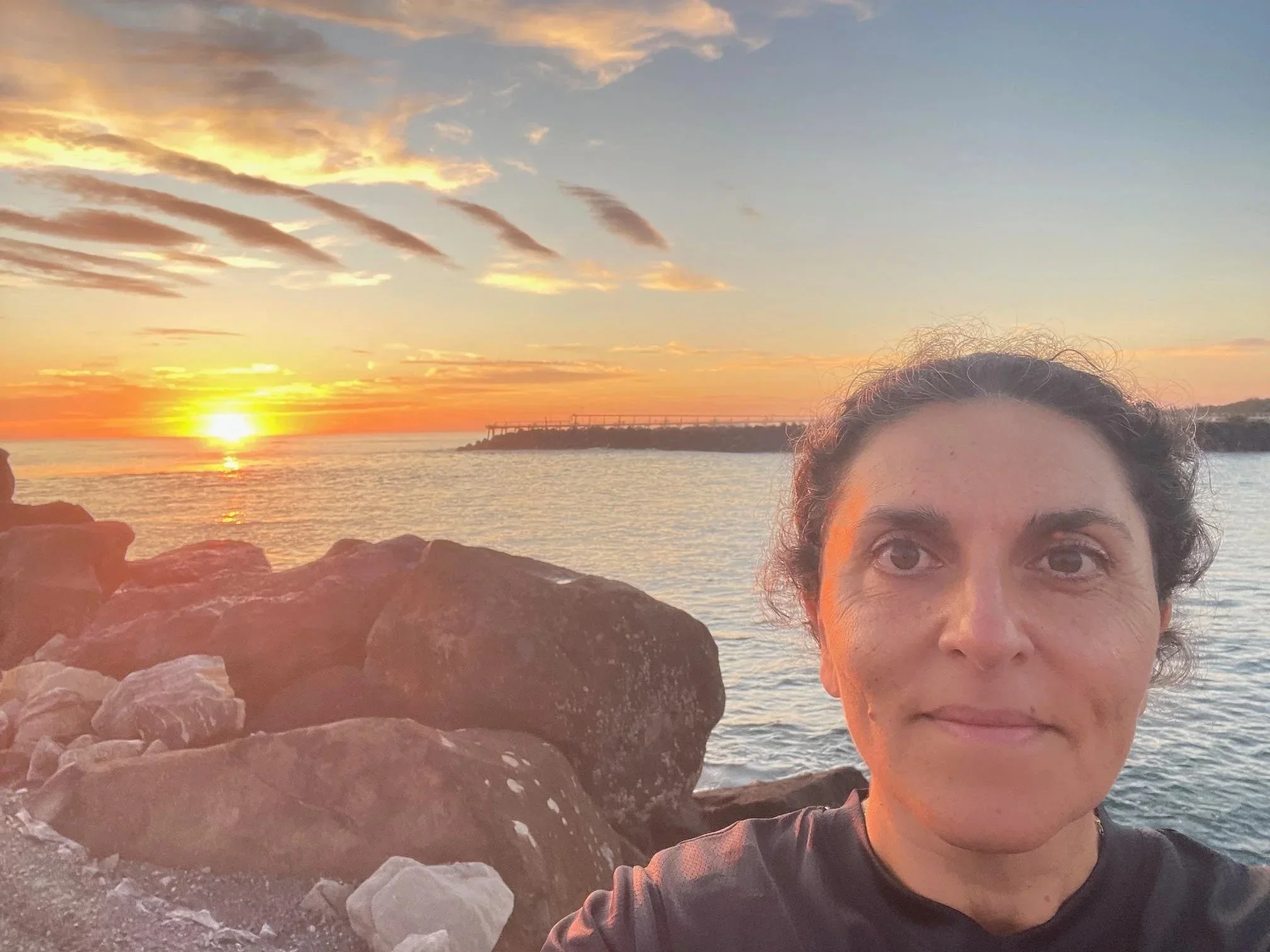 Woman sitting on rocks at the beach during sunset, wearing a black sleeveless shirt with flags and logos, with glasses on her head and a watch, smiling at the camera.