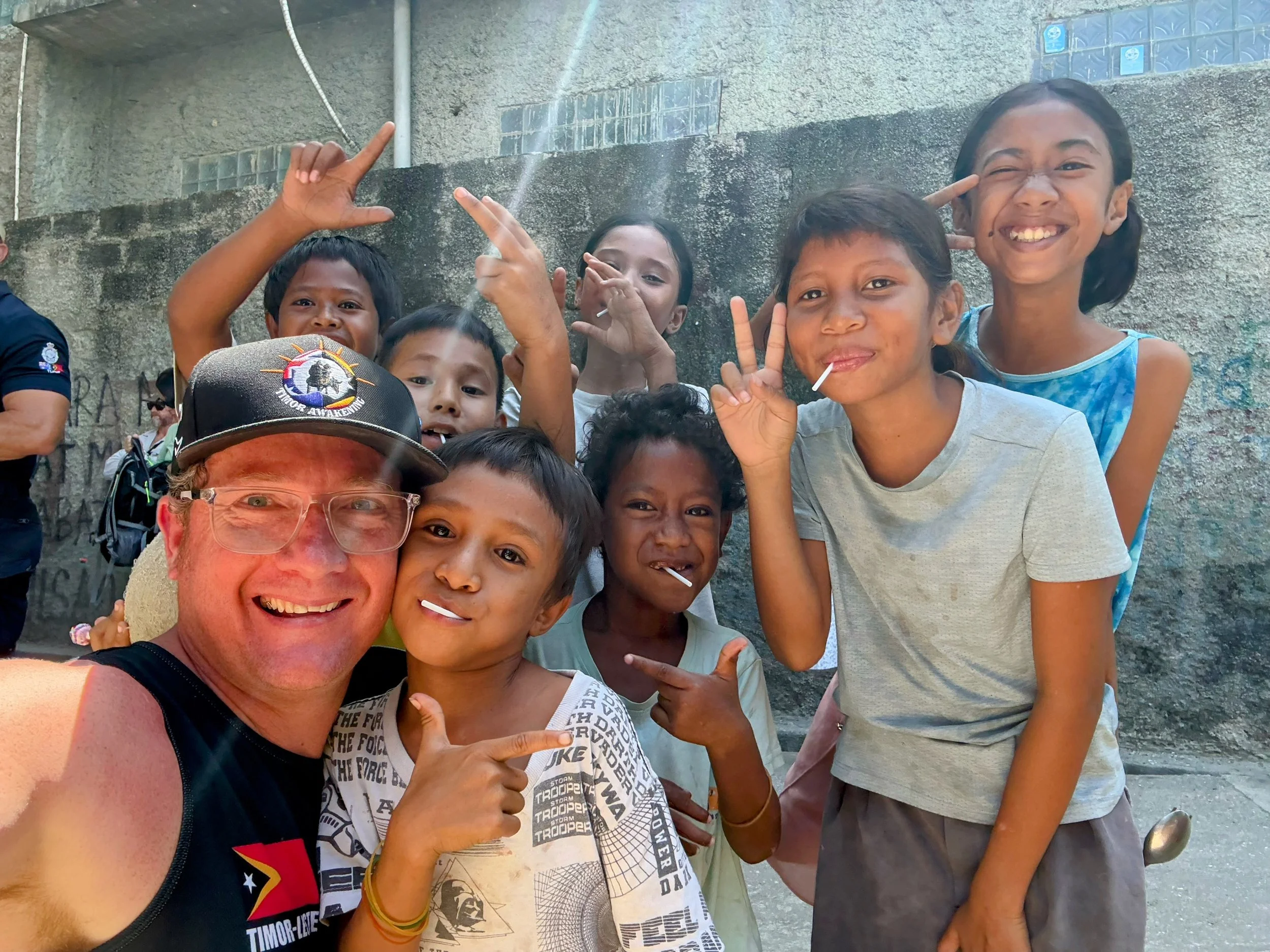A man wearing a dark blue suit, white shirt, and glasses standing against a plain gray background