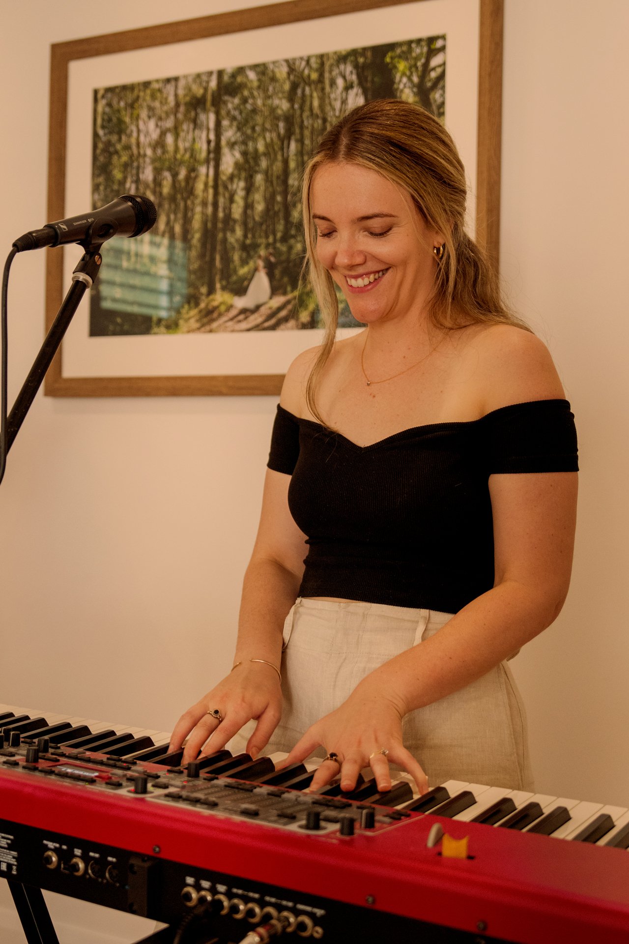 A woman playing a keyboard with a smile, wearing an off-the-shoulder black top and a beige skirt, in a room with a framed photo of a forest on the wall.