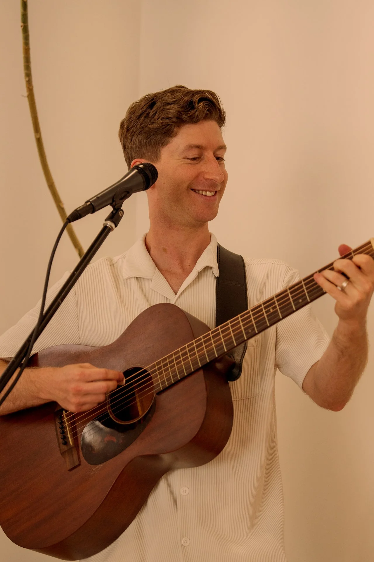 A man playing acoustic guitar and smiling, standing near a microphone.
