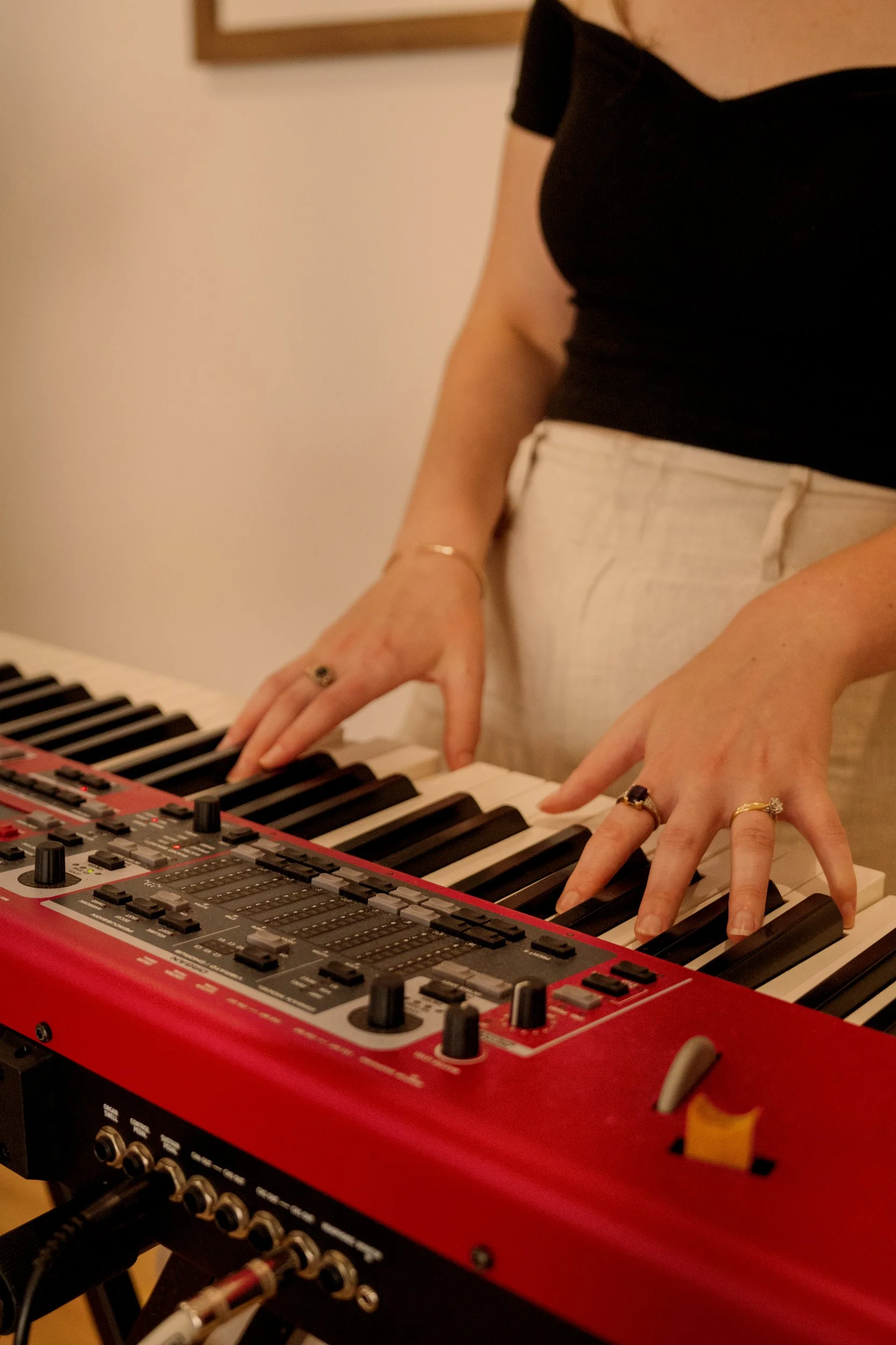 Close-up of a person playing a red synthesizer keyboard, with focus on their hands, wearing rings and a gold bracelet, in a cozy indoor setting.