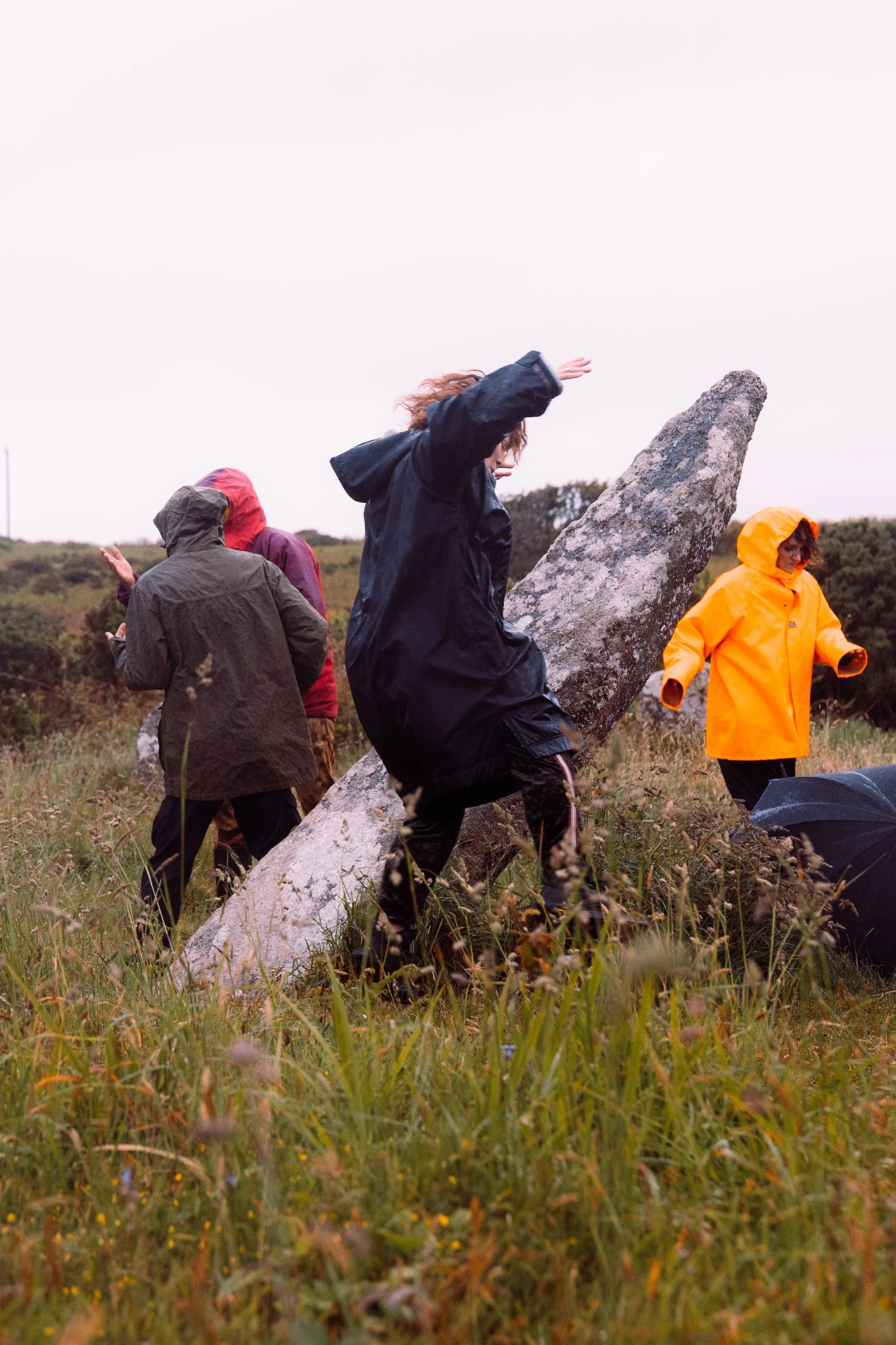 A middle aged white woman wearing a dry robe jumps as she passes a large standing stone. It is raining and there is long grass. In the background you can see other people in raincoats also dancing.