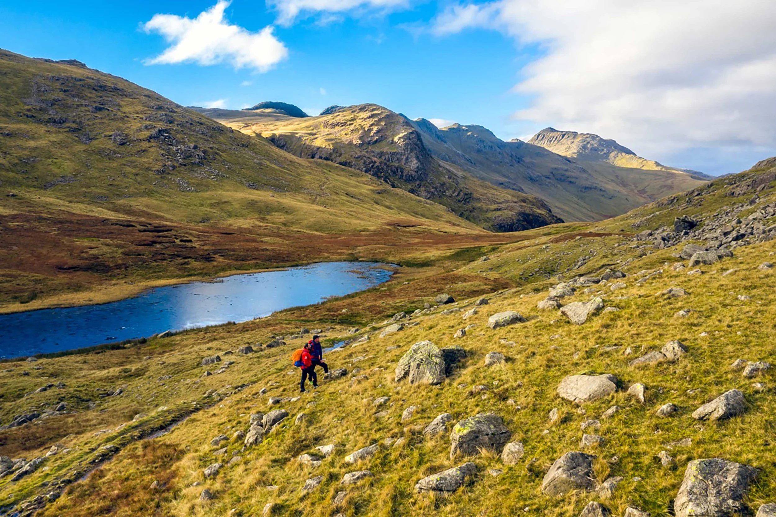 Hikers trekking along a grassy hillside with rocky terrain, a small lake, and distant mountains under a blue sky with clouds.
