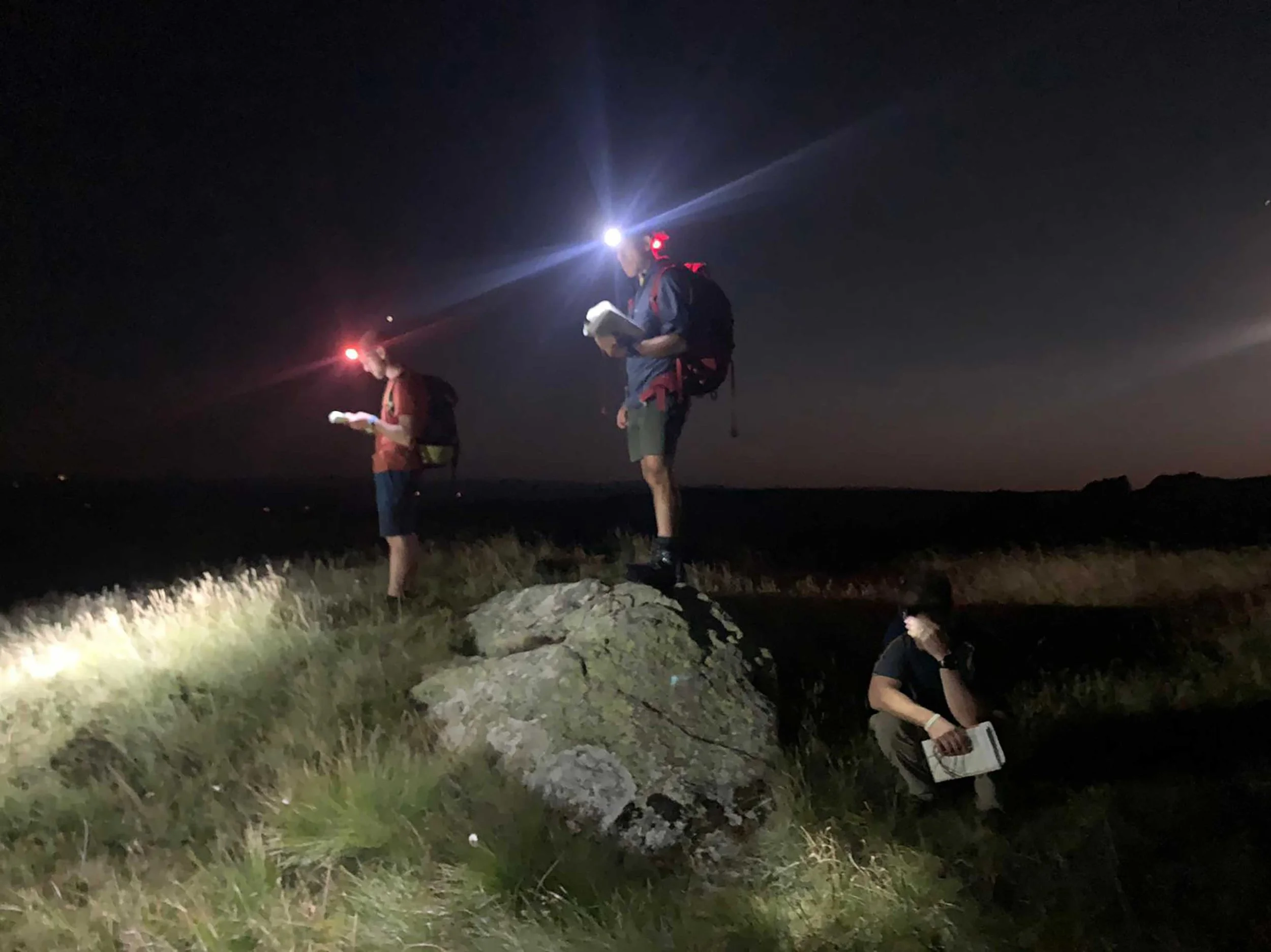 Three people with headlamps and backpacks hiking at night, consulting maps on a grassy area.