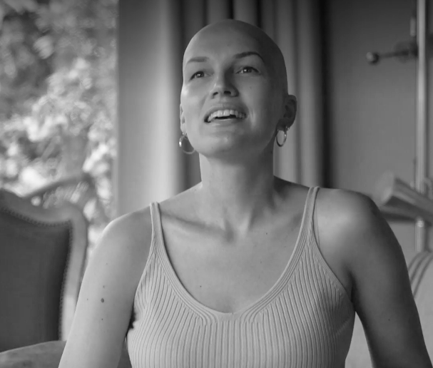 Black and white photo of a woman with a shaved head, wearing a sleeveless top, sitting indoors with curtains and a window in the background.