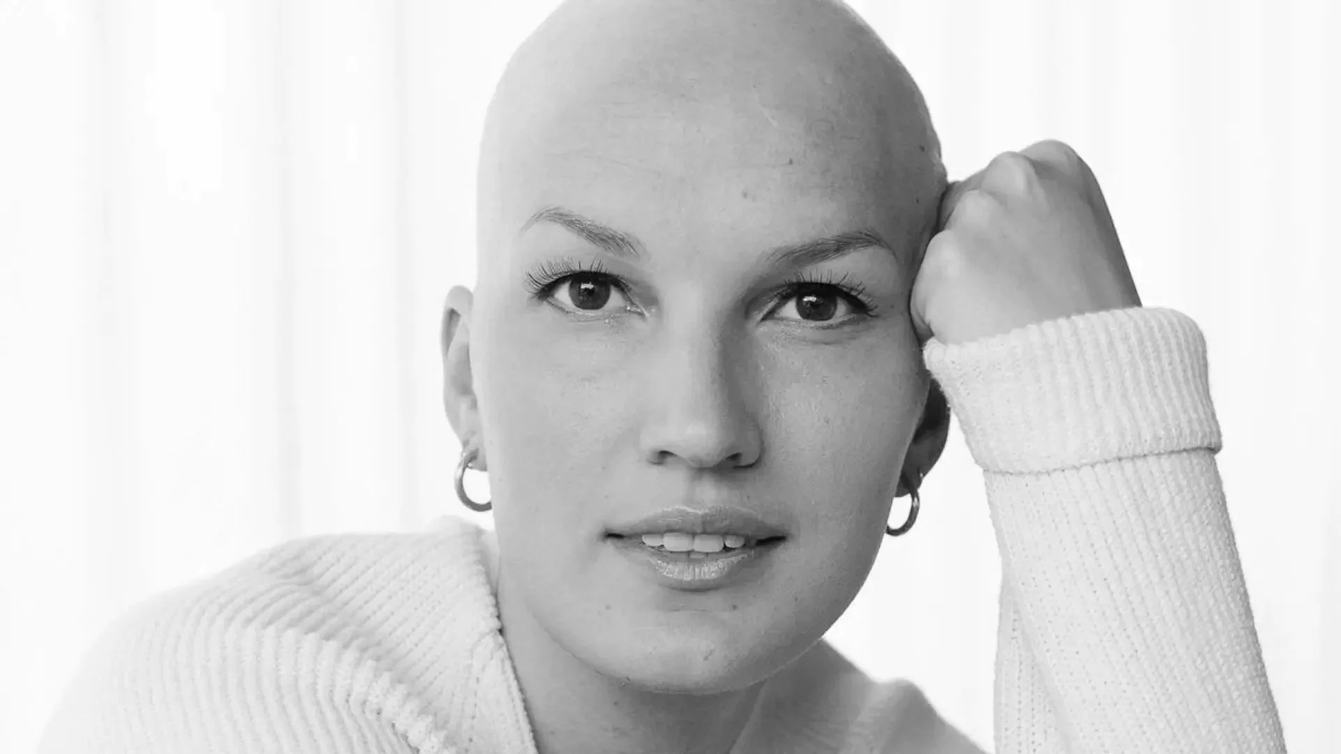 A black and white close-up of a woman with a shaved head, wearing a light-colored sweater, looking at the camera with a slight smile, resting her head on her hand.