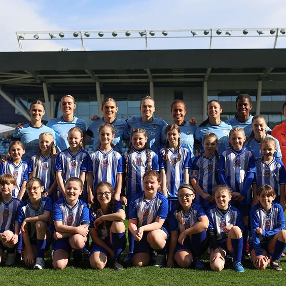 Group photo of a women's soccer team, including young girls and adult women, wearing blue and white striped uniforms, standing and sitting on a soccer field with a stadium in the background.