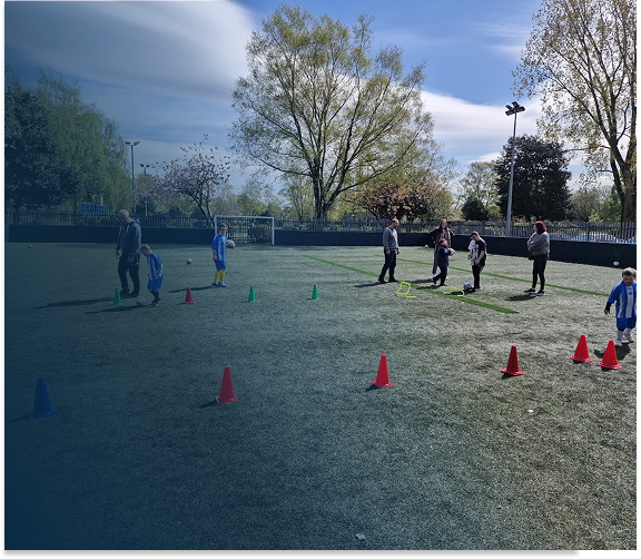 Children and adults participating in a soccer game or practice on a grass field, with orange, blue, and green cones set up for drills, and trees and a cloudy sky in the background.
