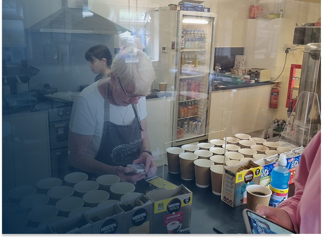 A woman with short gray hair taking an order at a coffee stand, with cups and drink ingredients on the counter, while a person with a pink jacket is holding a phone nearby. Behind the counter, there are two other people working in the kitchen area.