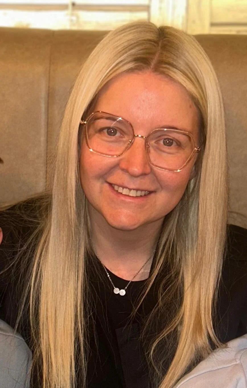 A woman with long blonde hair wearing glasses and a black shirt, smiling at the camera.